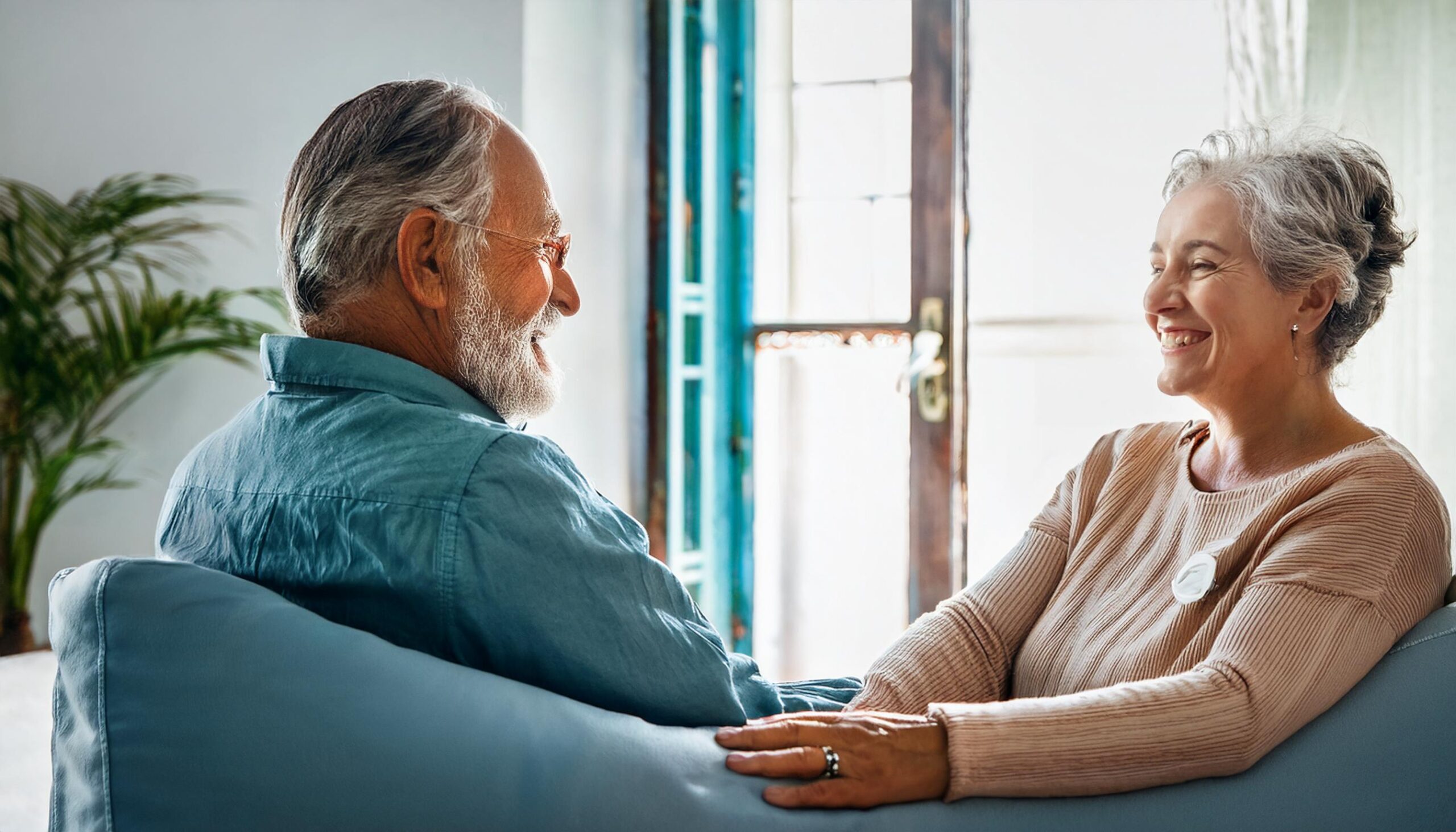 Una pareja de personas mayores sonríen mientras conversan en un ambiente hogareño iluminado por la luz natural, con plantas y una ventana al fondo