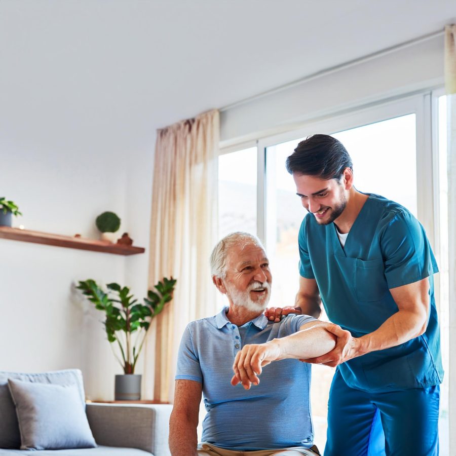 Un fisioterapeuta sonriente ayuda a un hombre mayor con ejercicios de movilidad en un ambiente hogareño iluminado y acogedor